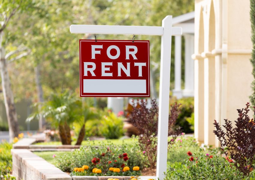 A red and white 'for rent' sign hangs from a white post in front of a rental home lawn.